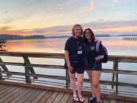 Two ladies on wooden pier in front of lake with sun setting in background