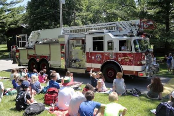 Fire Apparatus Parade and Appreciation Day attendees sit in the grass in front of fire truck