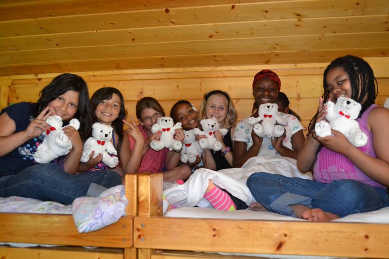 Young girls post on a bunkbed in their cabin, holding up matching stuffed teddy bears