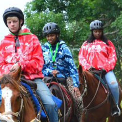 A group of young children wearing helmets and Camp "I am Me" sweatshirts riding horses through a green field.